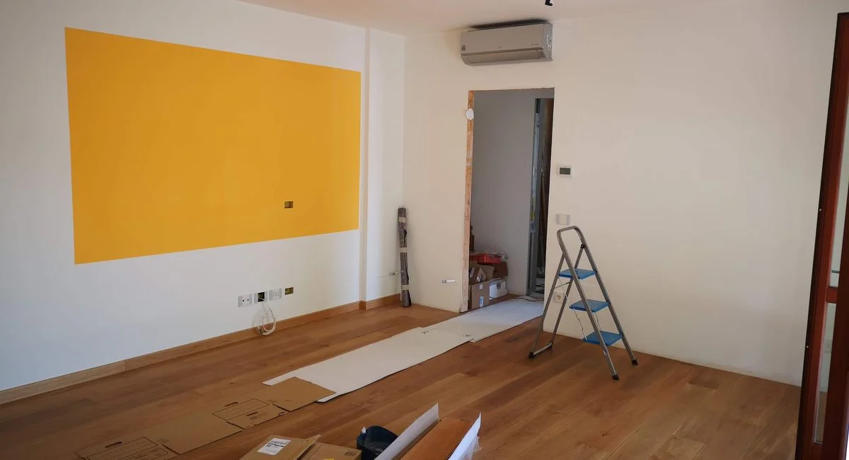 A bright, empty room under renovation with ladder, tools, and wooden flooring.