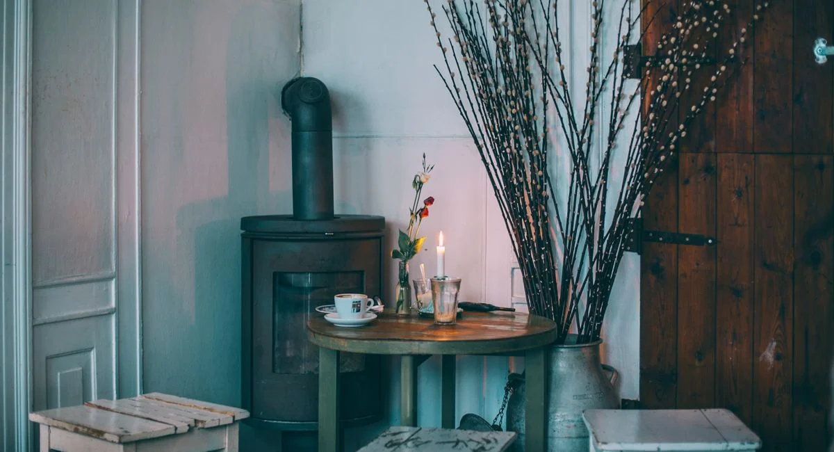 Interior of country house with shabby stools around table with served drinks near iron stove and dry branches in metal vase