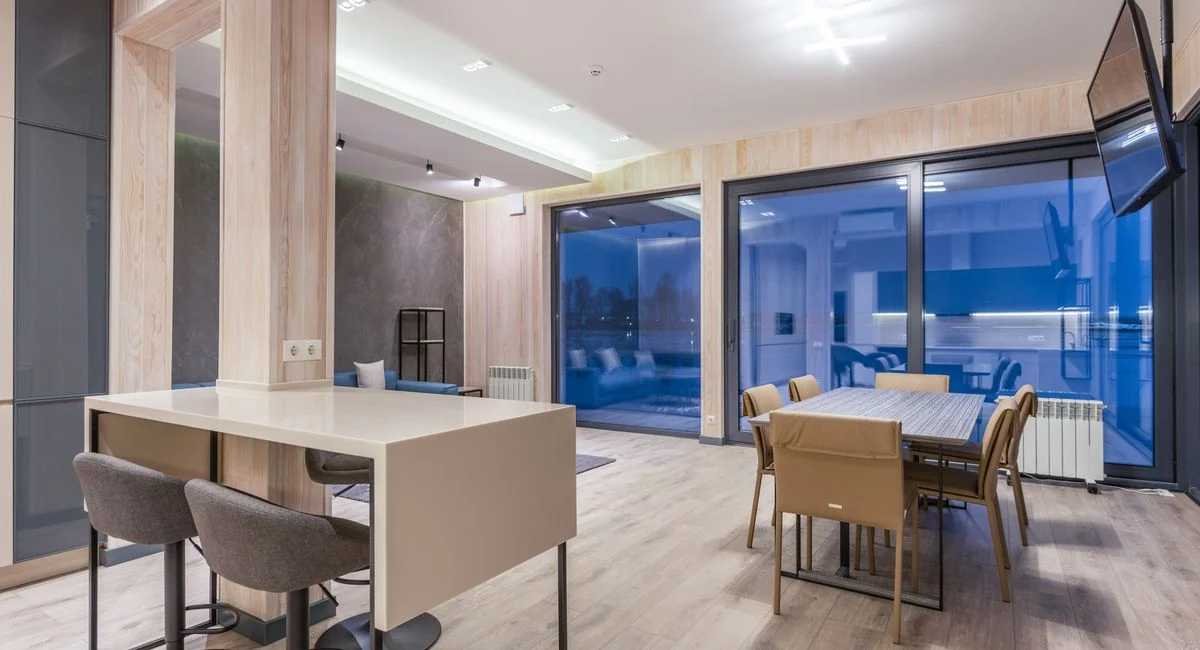 Dining tables with comfortable chairs in spacious apartment with wooden floor and walls and blue glass terrace doors