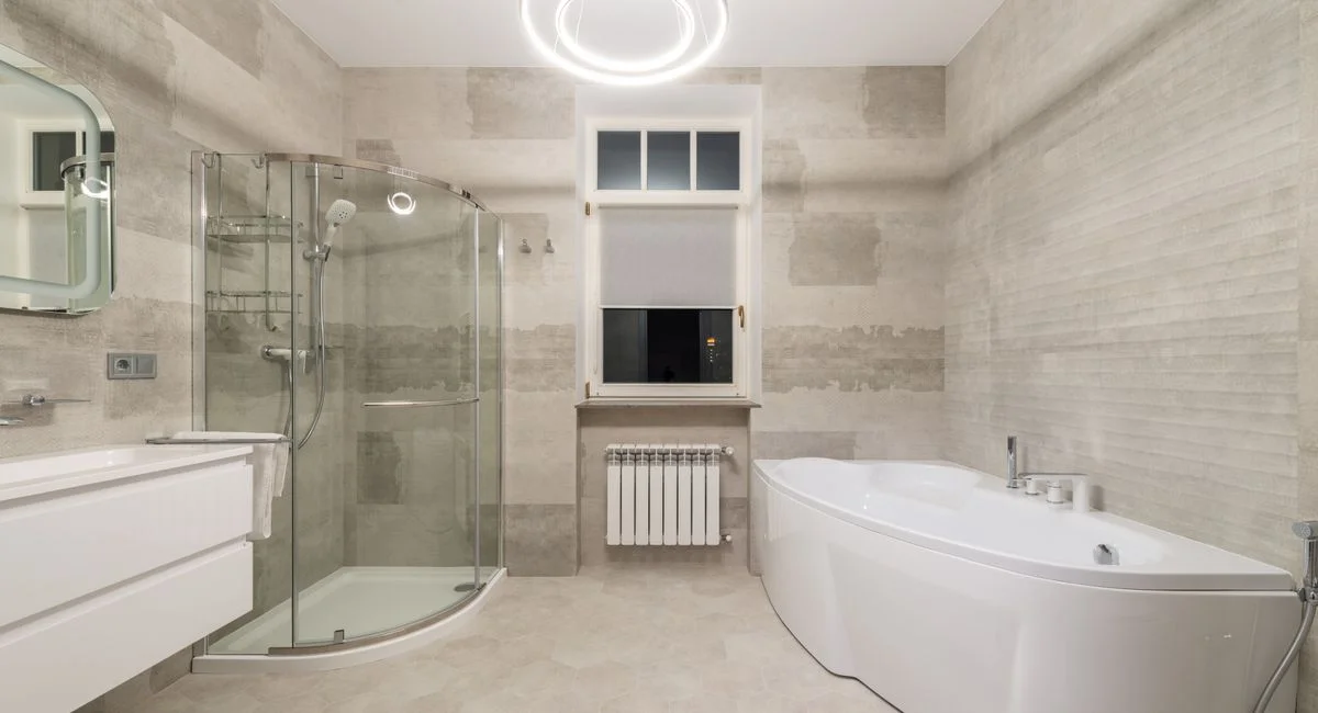 White bathtub near sink with mirror and glass shower cabin placed in stylish modern bathroom with window and glowing chandeli