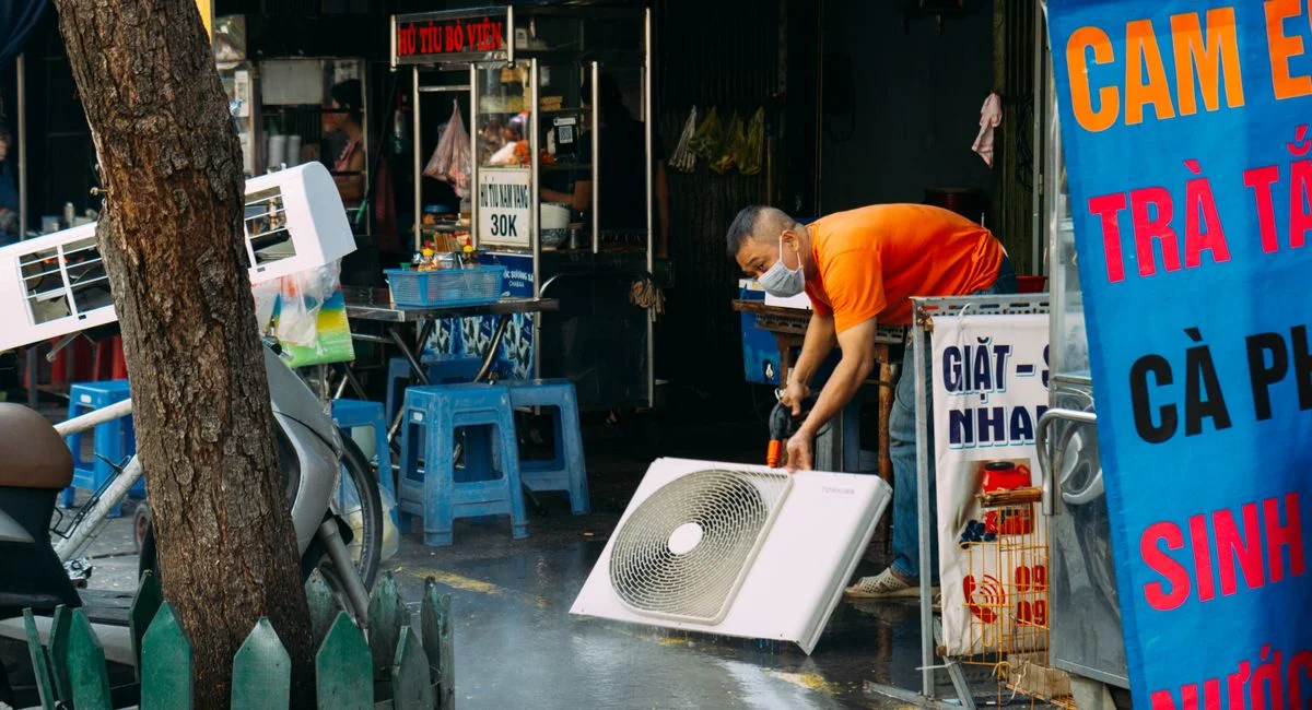 A man cleans a fan on a vibrant urban street with market stalls.