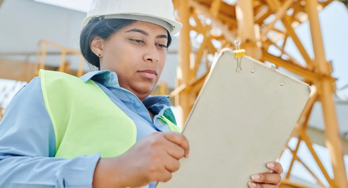 Confident female construction worker inspecting site with clipboard and helmet.