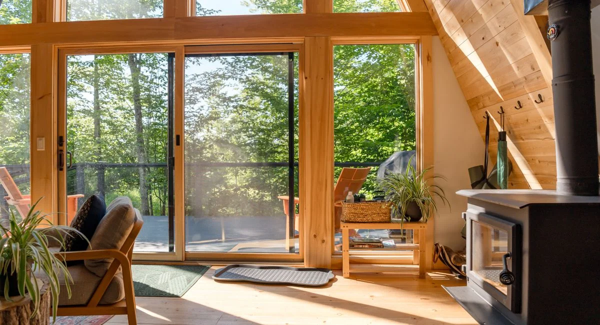 Sunlit interior of a cozy A-frame cabin with a wood stove, overlooking a green deck.