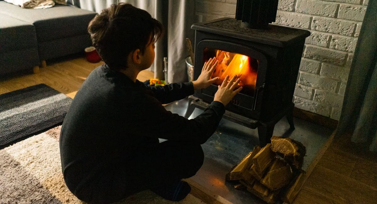 A child warms hands by a wood stove in a cozy living room, creating a warm winter ambiance.