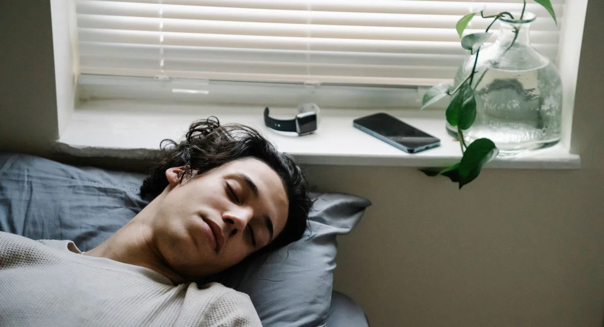 High angle of young ethnic male sleeping in bed with smartphone and smart watch placed near head on windowsill
